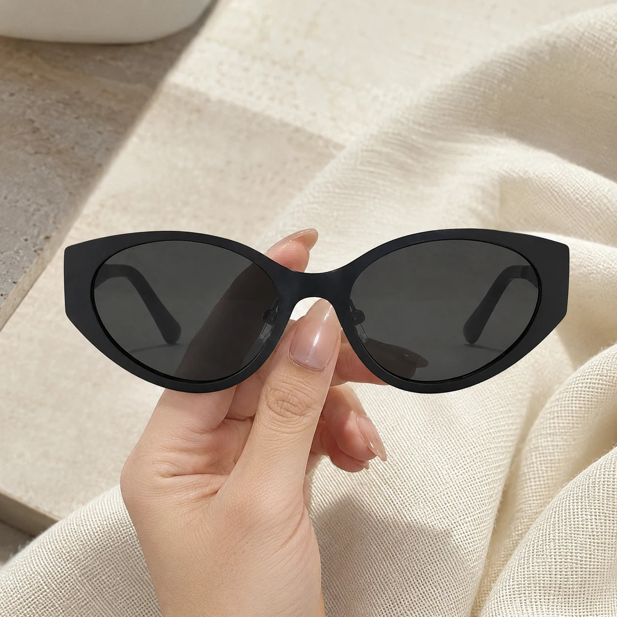A hand holding up the Lyra black cat-eye sunglasses with dark lenses against a soft cream fabric background.