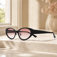 Lyra designer cat-eye sunglasses with thick black frames and gradient pink lenses displayed on a beige marble surface by a window