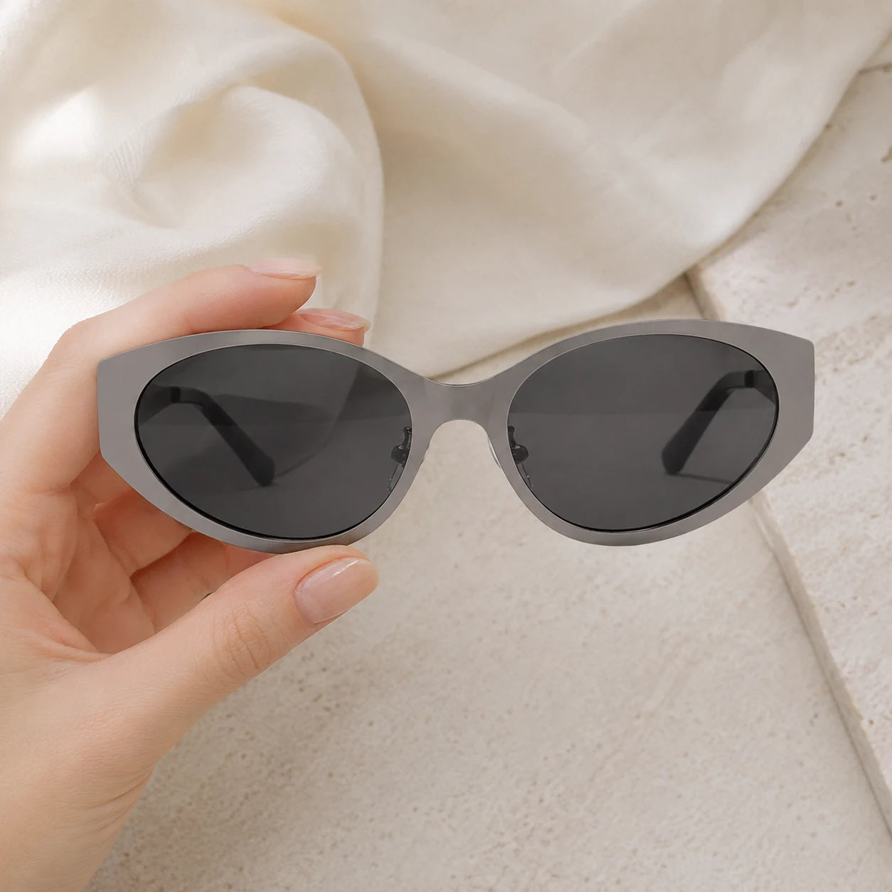 A hand holding up the Lyra stone gray cat-eye sunglasses against a soft white draped fabric background.