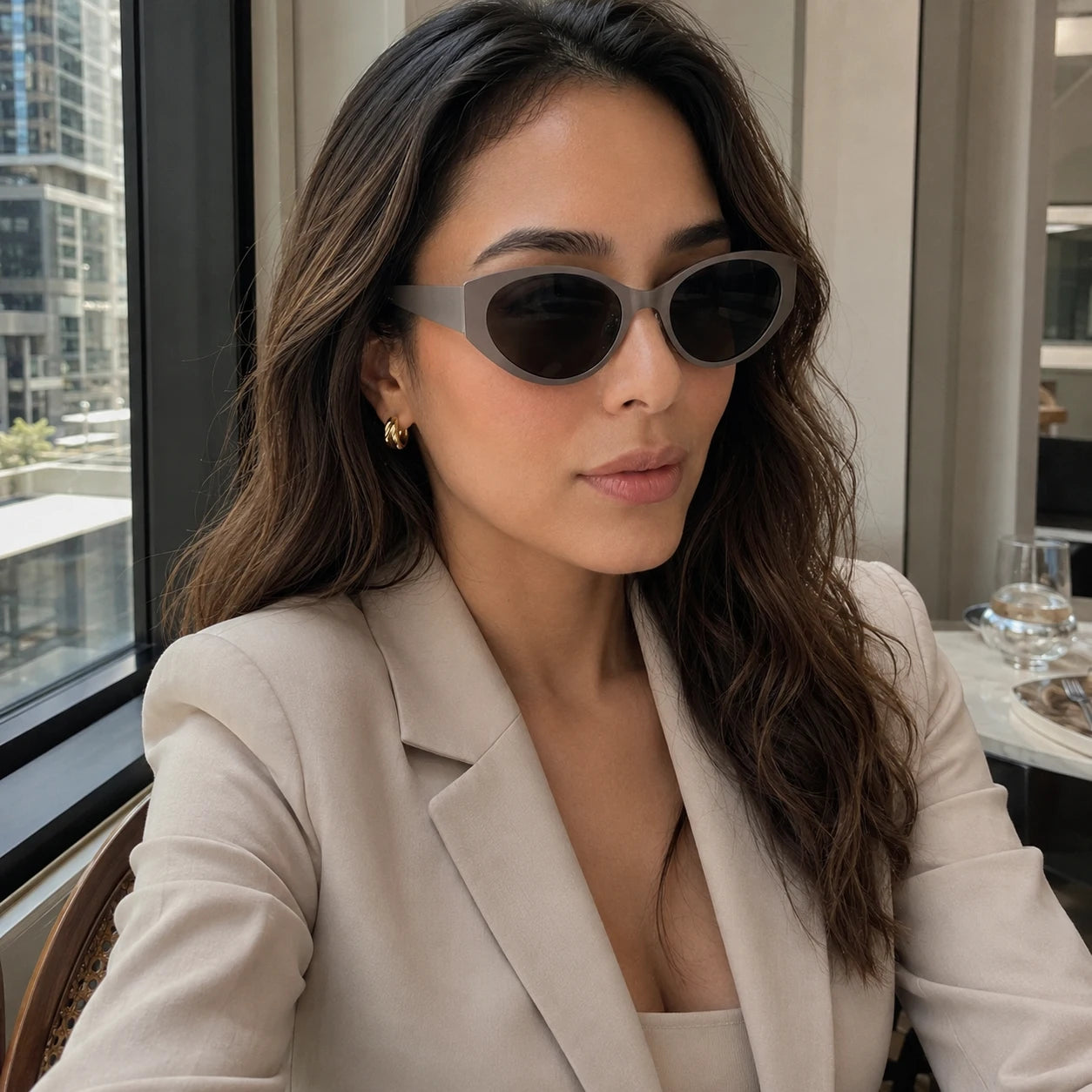 A stylish woman in a beige blazer wearing Lyra neutral gray retro sunglasses inside a modern venue.
