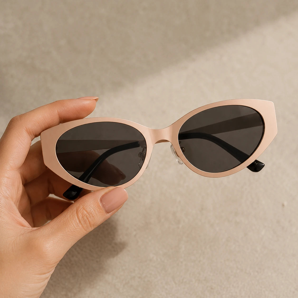A hand holding up the Lyra nude pink cat-eye sunglasses against a neutral sandy textured background.