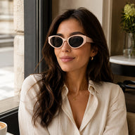 A chic woman wearing Lyra nude pink retro sunglasses while sitting by a window at a coffee shop.